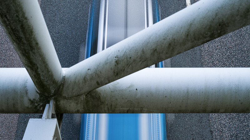Two weathered concrete pipes intersecting diagonally in front of a textured gray wall and blue metal frame.