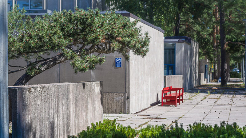 Modern concrete building with blue door next to a gnarled tree, red chair on paved courtyard.