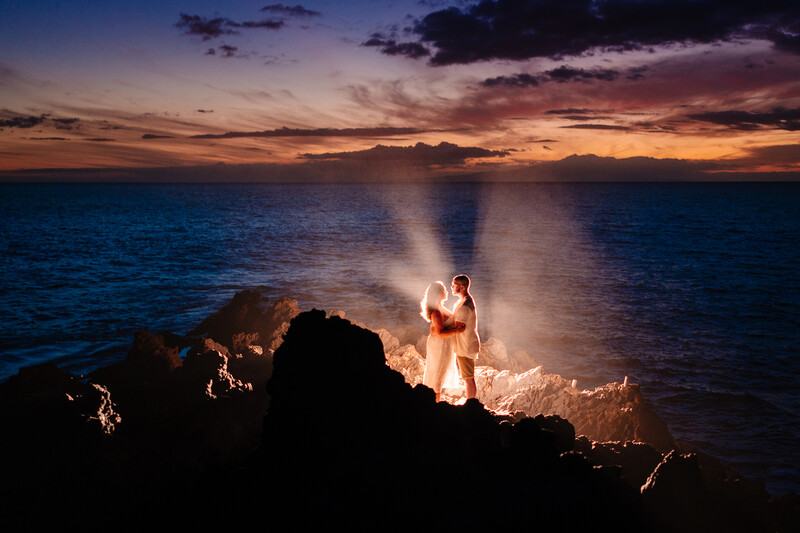Couple in white clothing standing on dark rocks by the ocean at twilight, illuminated by golden light rays from the sunset sky.