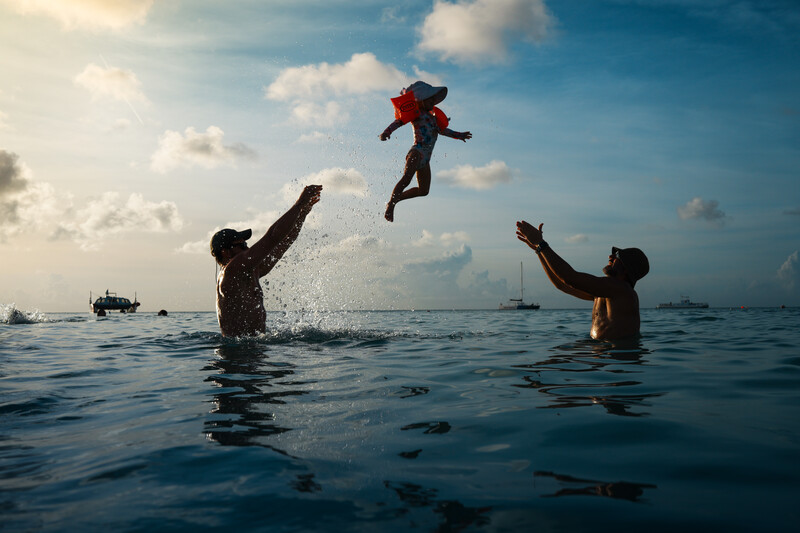 Two adults in water toss a child in a red outfit into the air against a blue sky with white clouds.