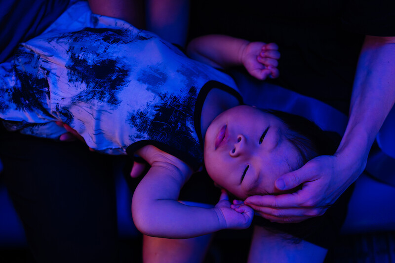 Sleeping infant cradled in adult hands under blue and purple gel lighting.