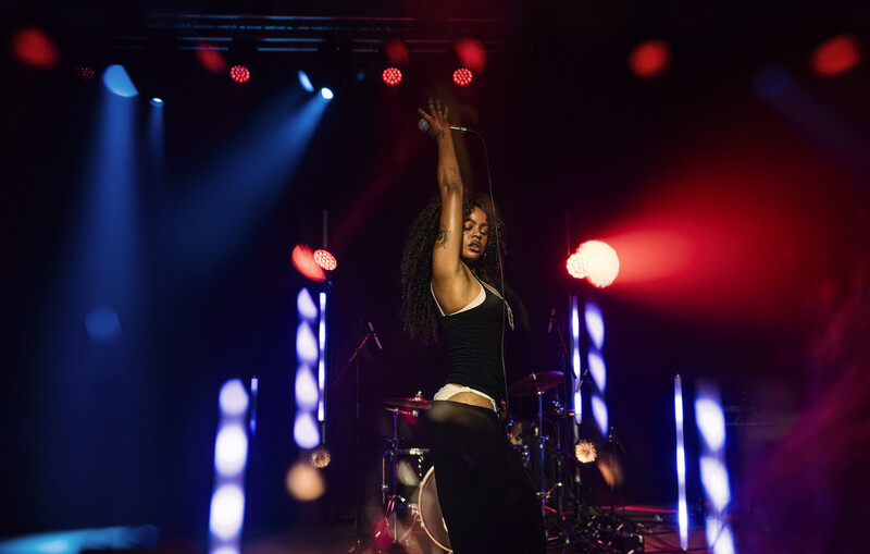Female performer on stage with arm raised, surrounded by blue and red concert lighting and illuminated vertical elements.