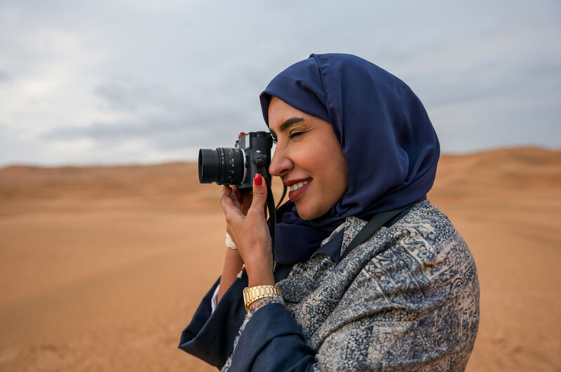 Woman in navy hijab and patterned jacket holding vintage film camera to her face in desert landscape.