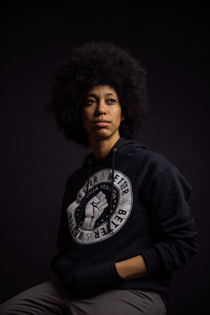 Person wearing black graphic hoodie against dark background, seated and looking toward camera.