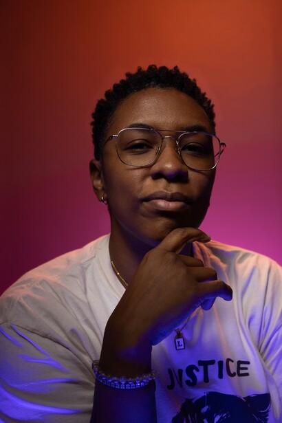 Young person wearing glasses and a light-colored shirt with blue text, posed thoughtfully against a gradient background.