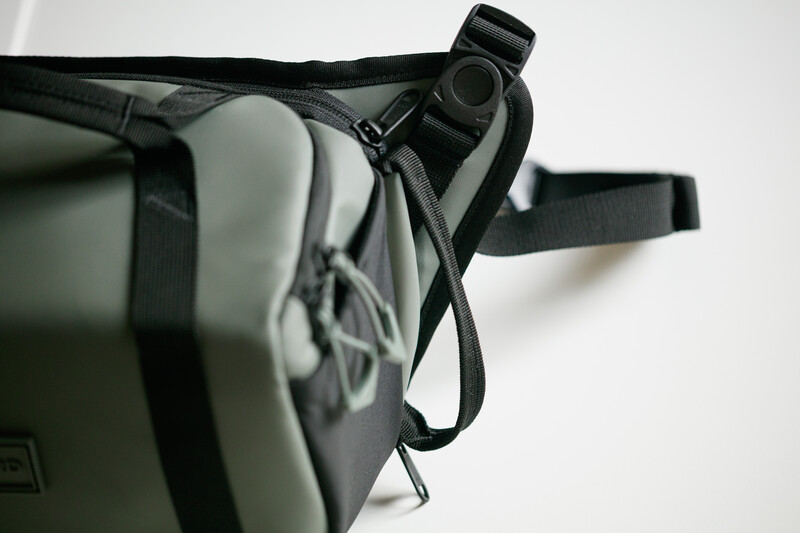 Close-up of a shoulder camera strap with attached small black binocular or spotting scope.