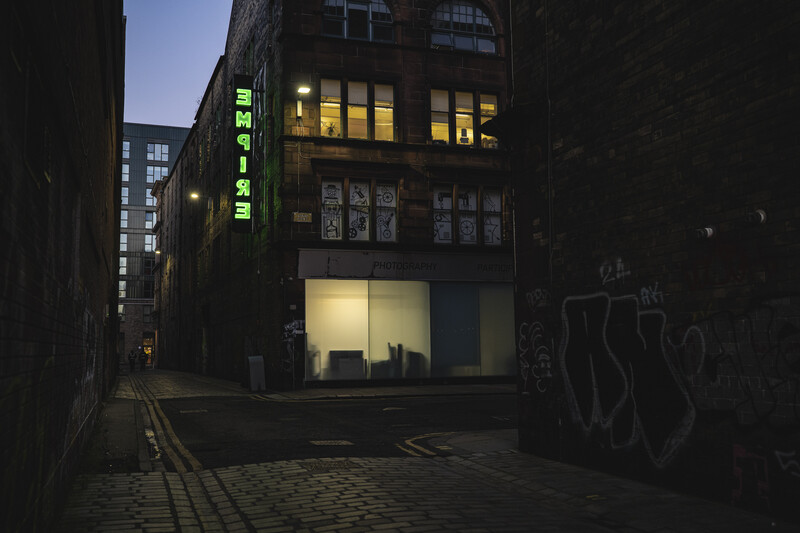 Illuminated storefront on a dark urban street lined with graffitied brick walls at dusk.