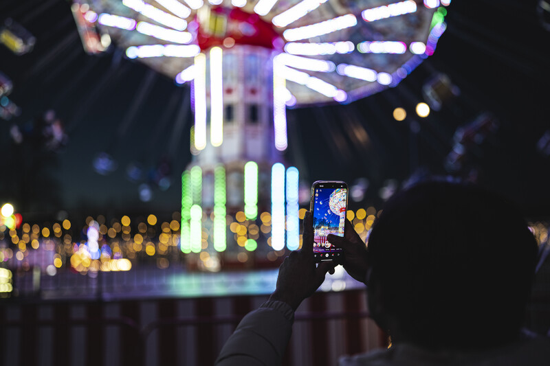 Person holding smartphone photographing illuminated Ferris wheel at night.