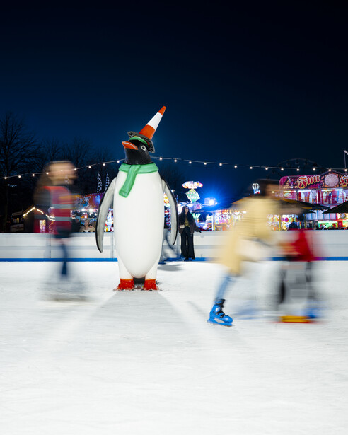 Penguin figure wearing a party hat on an ice rink at dusk with blurred skaters moving around it.