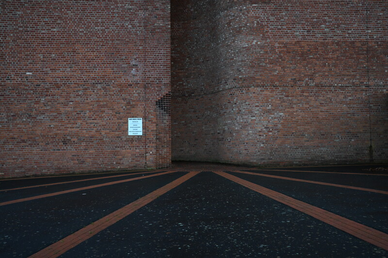 Symmetrical perspective view down a narrow corridor formed by two brick walls, with converging copper or rust-colored lines on dark pavement.