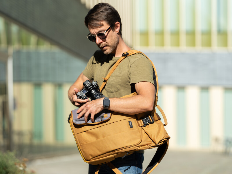 Photographer wearing sunglasses adjusts camera settings while holding a mustard-colored camera bag.
