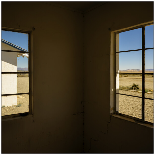 Dark interior corner with two windows framing expansive flat landscape views under clear sky.
