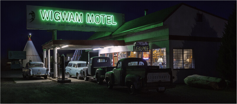 Vintage motel at night with neon sign, parked classic cars, and illuminated storefront.