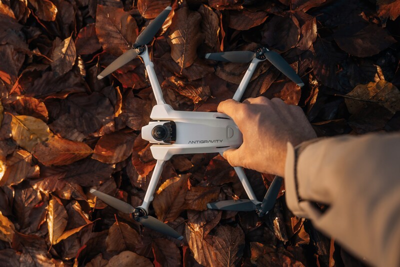 Hand holding a white compact drone with foldable arms over a bed of dried brown leaves.