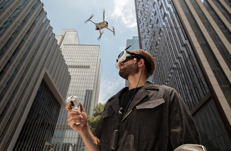 Drone pilot holding controller while looking up at flying quadcopter between tall city buildings.