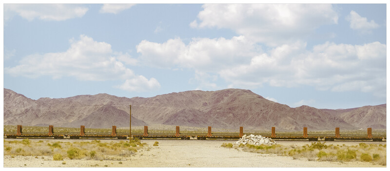 Rusted railroad tracks stretching across a sparse desert landscape with mountains in the background.