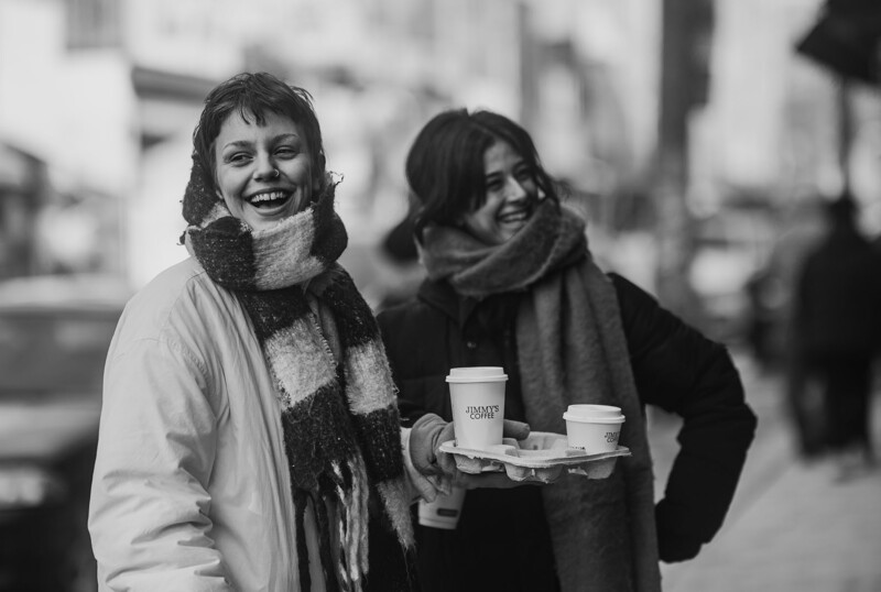 Two women holding coffee cups on a city street, smiling at the camera in black and white.