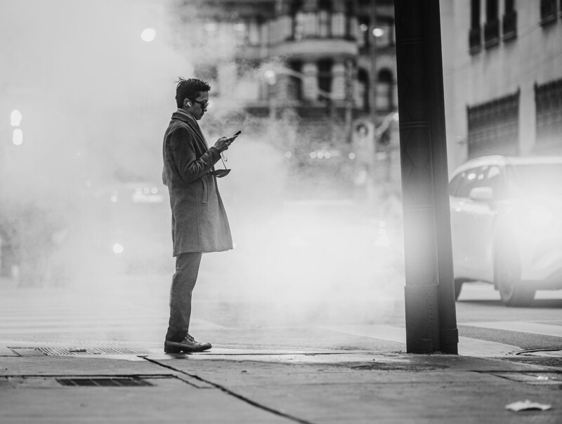 Man in coat standing on urban street checking phone amid heavy fog and building silhouettes.