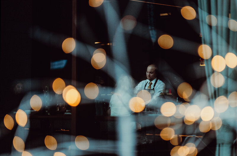 Man in light blue shirt seated by window with warm bokeh lights in foreground and background.