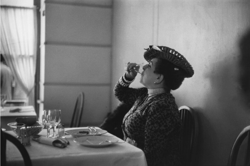 Woman in formal attire seated at a dining table, adjusting her decorative hat while looking upward.