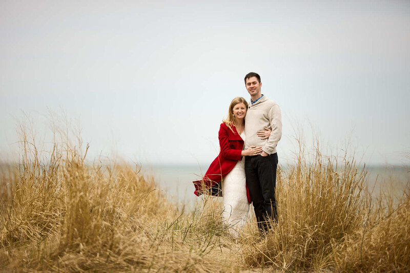 Couple posing together in dried beach grass with overcast sky and water in background.
