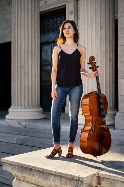 Musician standing with cello on classical stone platform with columns behind her.