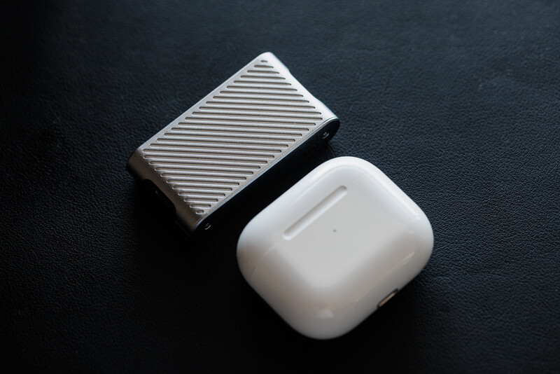 White wireless earbuds case and silver textured portable hard drive on dark surface.