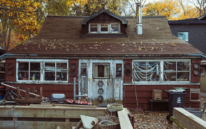 Weathered red cottage with boarded windows and deteriorating exterior surrounded by autumn trees.