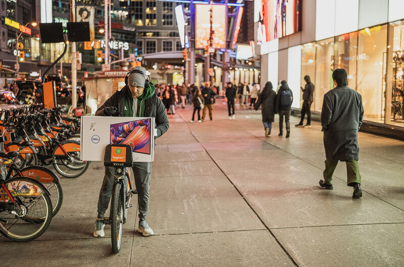 Cyclist carrying a white package rides through a crowded urban plaza at dusk.