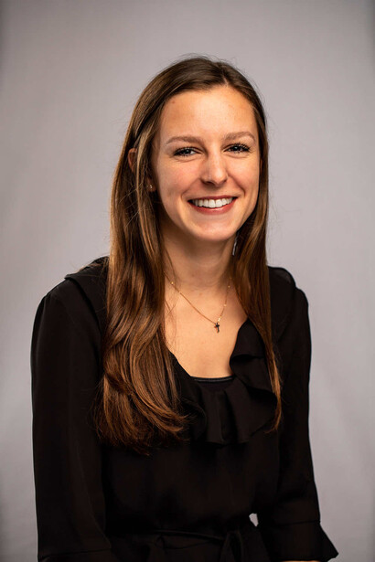 Woman with long brown hair wearing a black blazer and necklace, smiling at camera against neutral gray background.