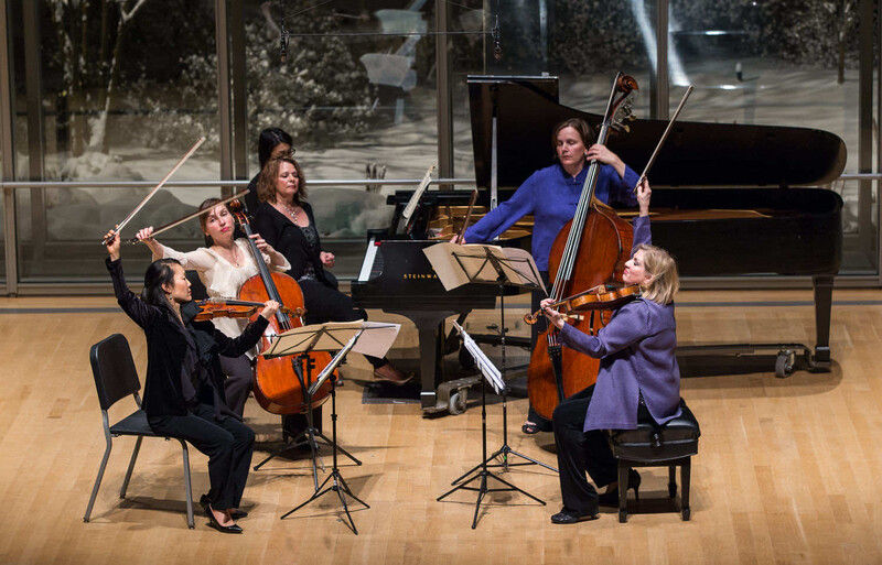 Five musicians performing chamber music with string instruments and piano in a concert hall.