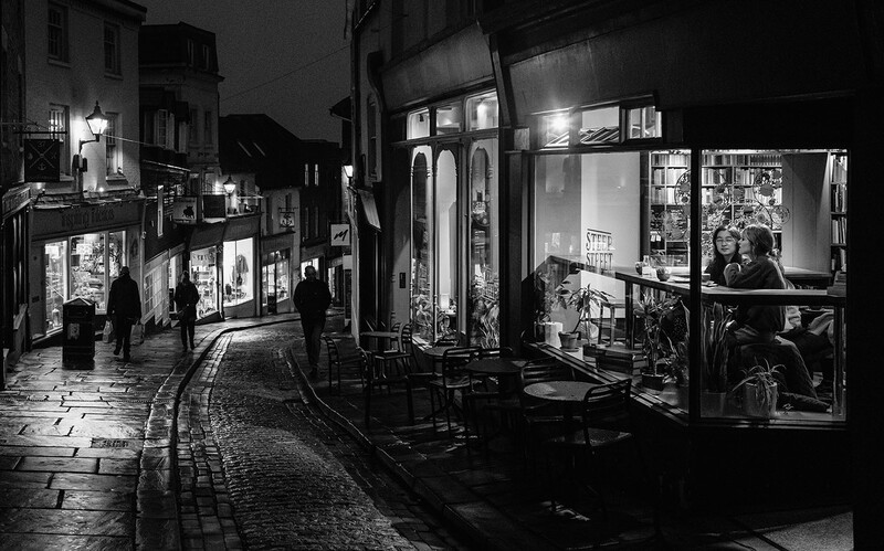 Nighttime street scene with illuminated storefronts and pedestrians on a wet cobblestone street.