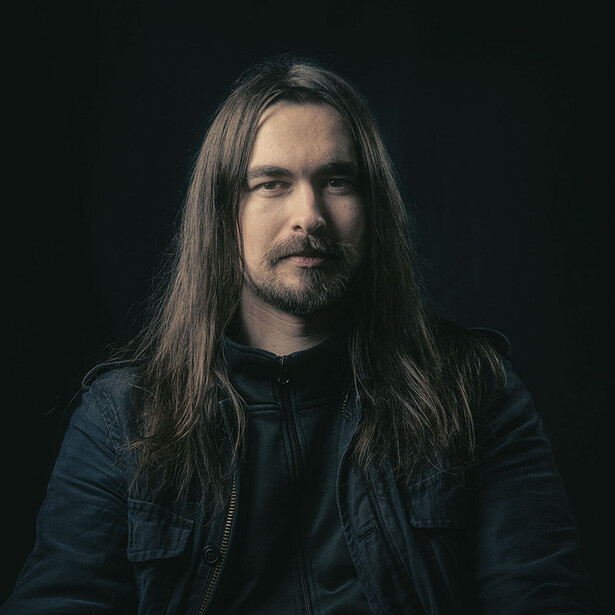 Man with long dark hair wearing a dark denim shirt against a black background.