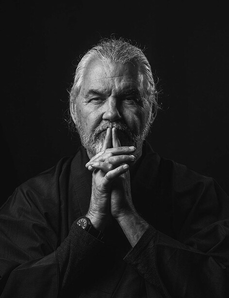 Bearded man in dark shirt with contemplative expression, hand near face against black background.