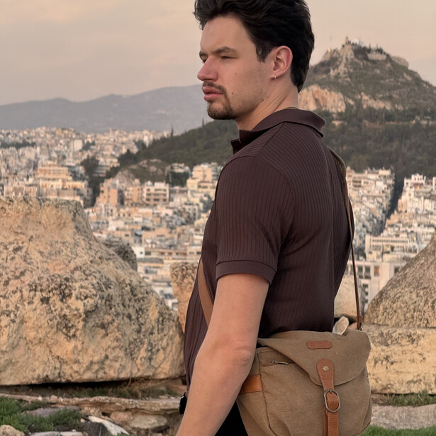 Man in brown shirt with canvas bag overlooking dense hillside cityscape at golden hour.