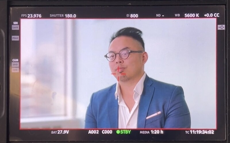 Screenshot of camera monitor displaying portrait of man in blue blazer and glasses during video production.