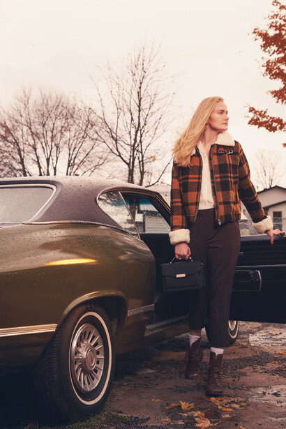 Woman in plaid shirt leaning against vintage car in autumn setting.