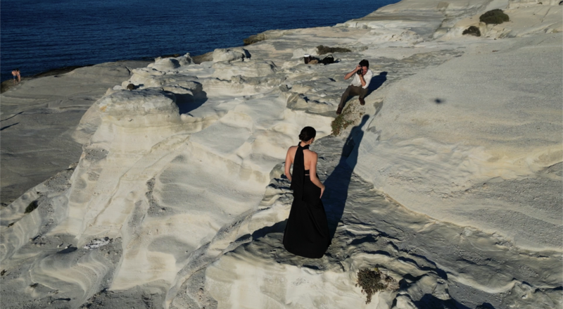 Woman in black dress posing on white rocky terrain overlooking blue water.