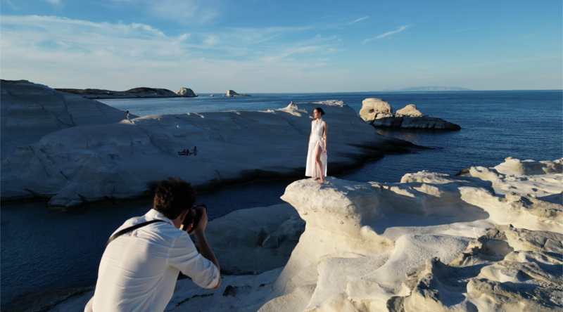 Photographer shooting a woman in a white dress posed on white rock formations overlooking the sea.