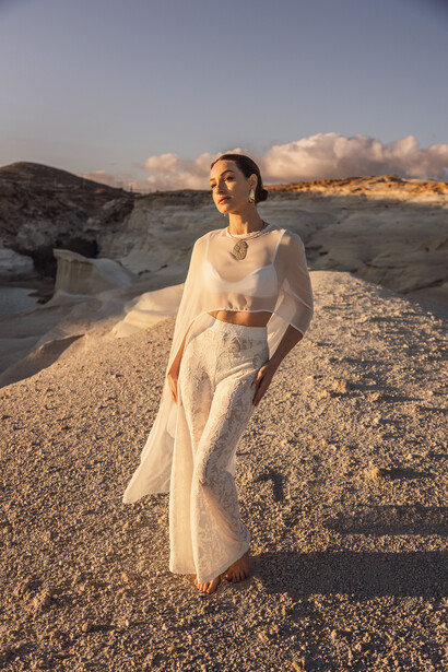 Woman in cream linen dress standing on rocky terrain during golden hour with mountains and clouds in background.
