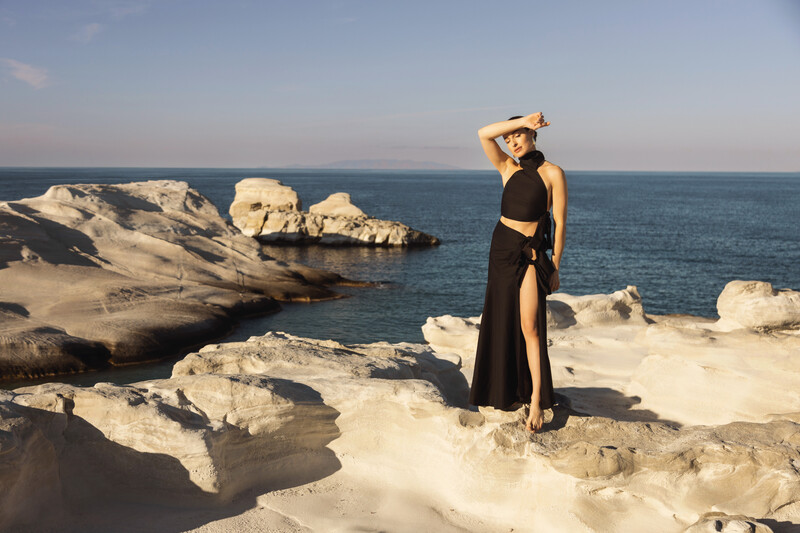 Woman in black dress and hat standing on white rocky outcropping overlooking the sea.
