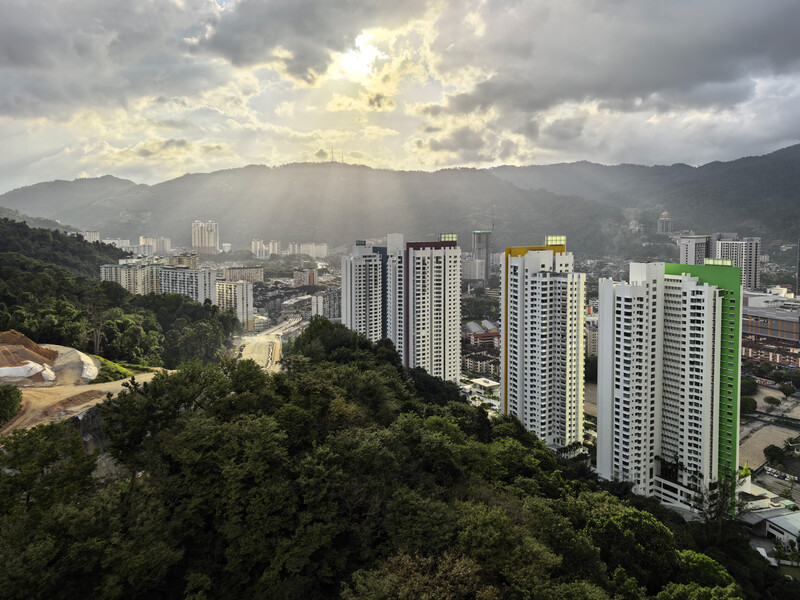 Aerial view of modern high-rise residential buildings with forested hills and distant mountains under dramatic cloudy sky.