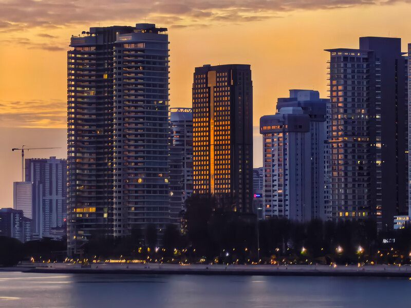 Urban skyline at sunset with illuminated high-rise buildings reflected in calm water.