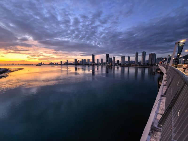 Cityscape reflected in still water at sunset, viewed from a pier with dramatic cloudy sky.
