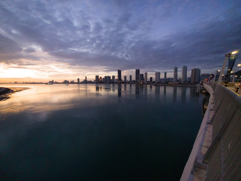Skyline cityscape reflected in calm water during twilight, photographed from a pier or waterfront structure.