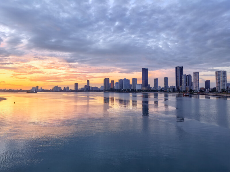 Urban skyline reflected in calm water at sunrise, with dramatic cloudy sky overhead.