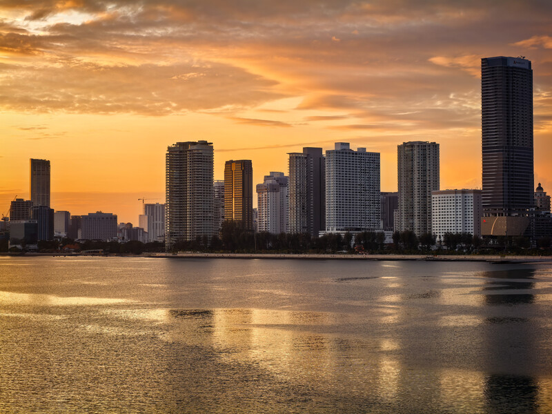 Urban skyline silhouetted against golden sunset sky, reflected in calm water below.