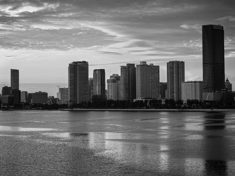 Monochrome cityscape with high-rise buildings reflected in still water under dramatic cloudy sky.