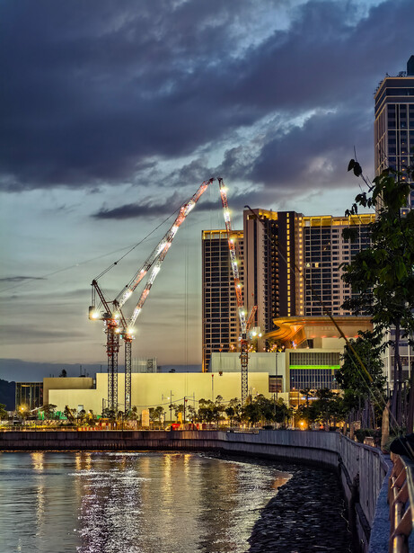 Illuminated high-rise buildings reflected in calm water at twilight with dramatic storm clouds overhead.