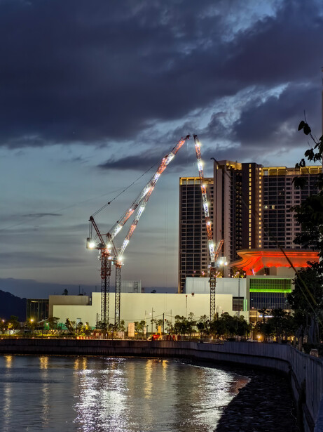Illuminated construction crane against dramatic twilight sky over waterfront buildings.